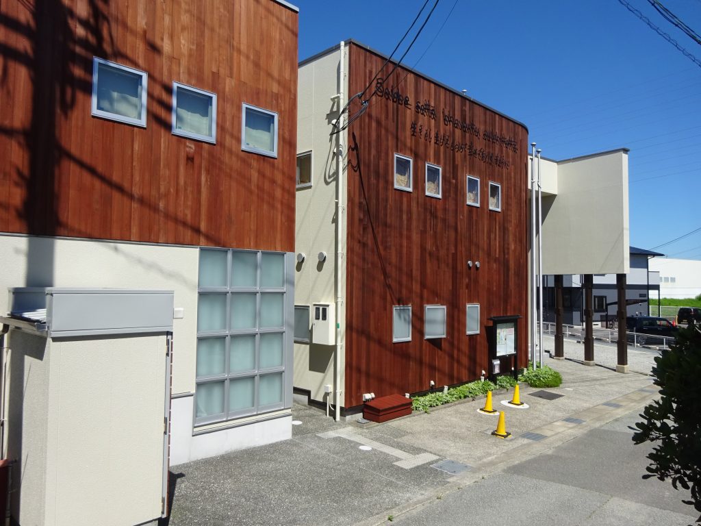 Modern building exterior with red wood siding and beige walls, row of square windows under a clear blue sky, cones in the parking area.