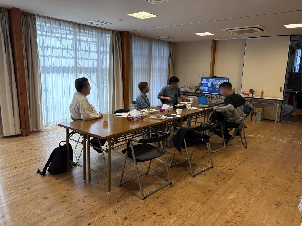 Conference room with several people seated around a long table, participating in a video conference shown on a monitor, with laptops, cups, and snacks on the table.