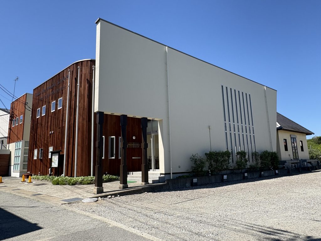 Modern building with a wood-paneled left section and light gray right section under a clear blue sky, with an entrance and vertical columns.