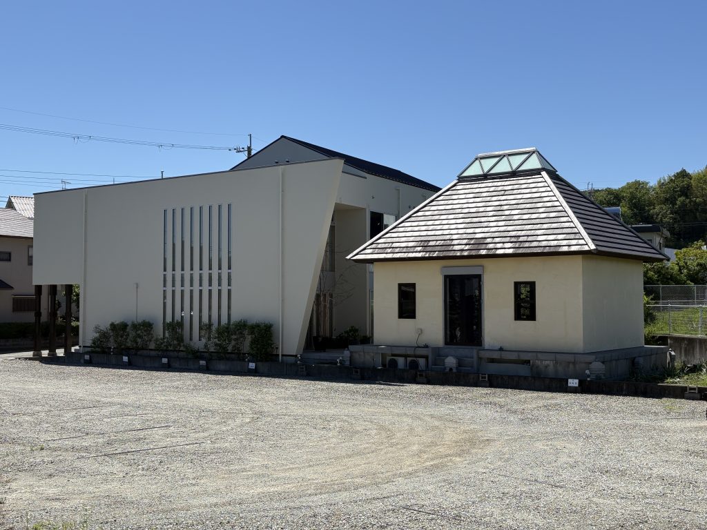 Modern white building with a sloped roof and a glass pyramid-like skylight on the right, set in a gravel lot under a clear blue sky.