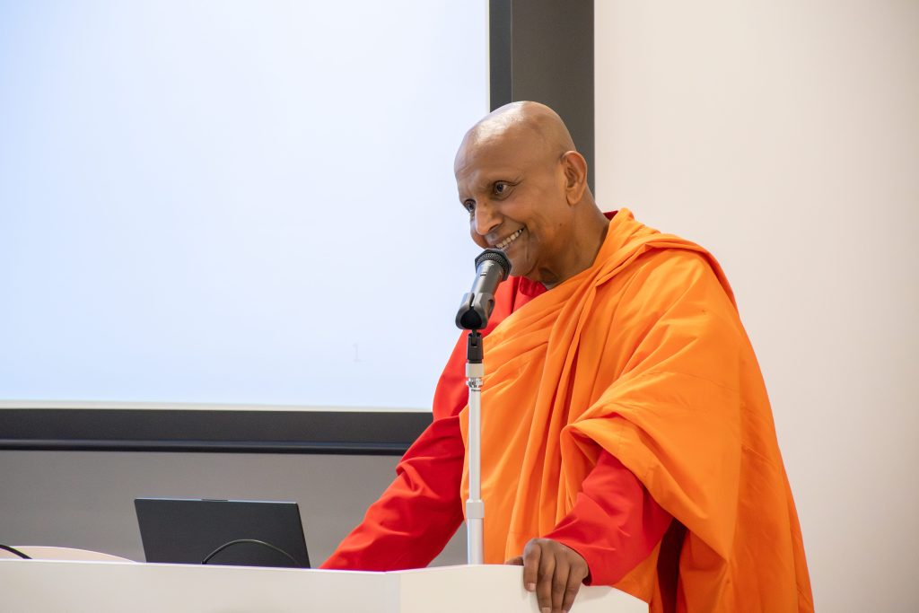 Buddhist monk in orange robes speaks into a microphone at a podium with a laptop nearby.