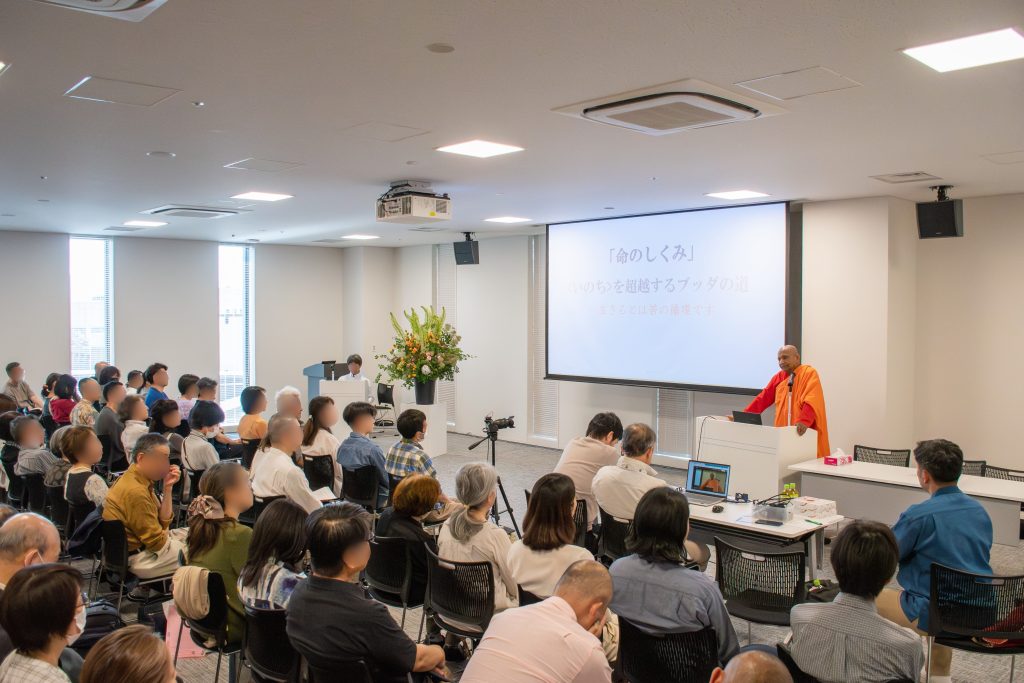 Audience seated in a conference room watching a speaker in orange robes at a podium, with a projection screen showing Japanese text nearby.