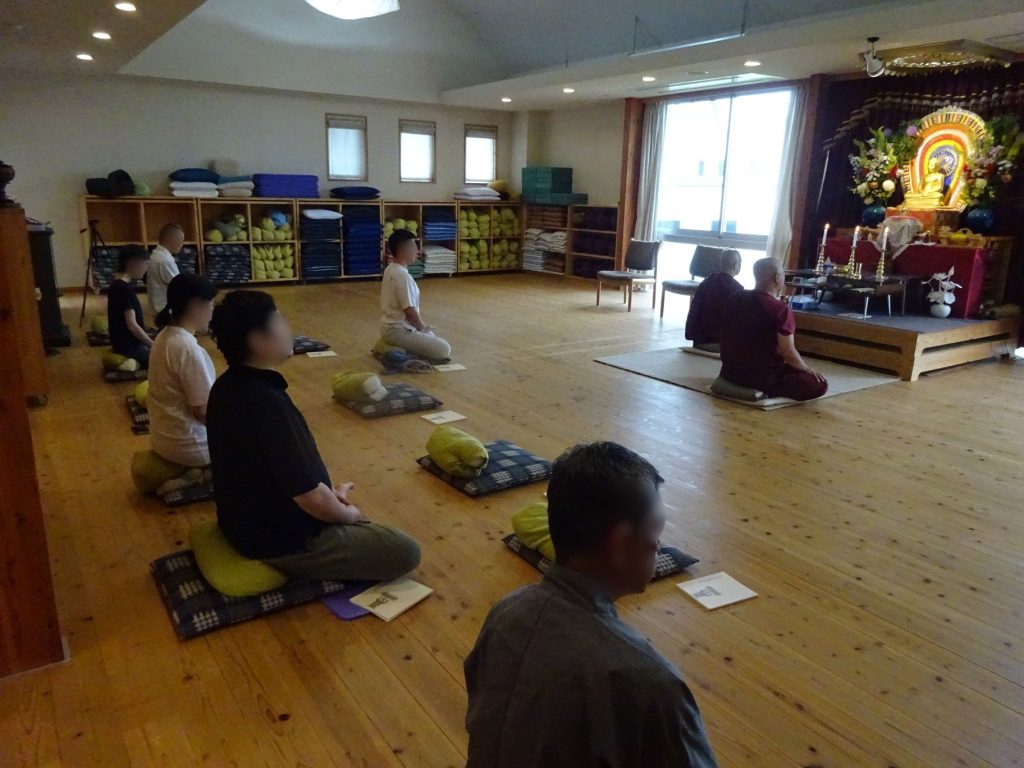 Group of people kneeling on cushions in a studio, facing a Buddha altar with flowers and candles in the front corner of the room.