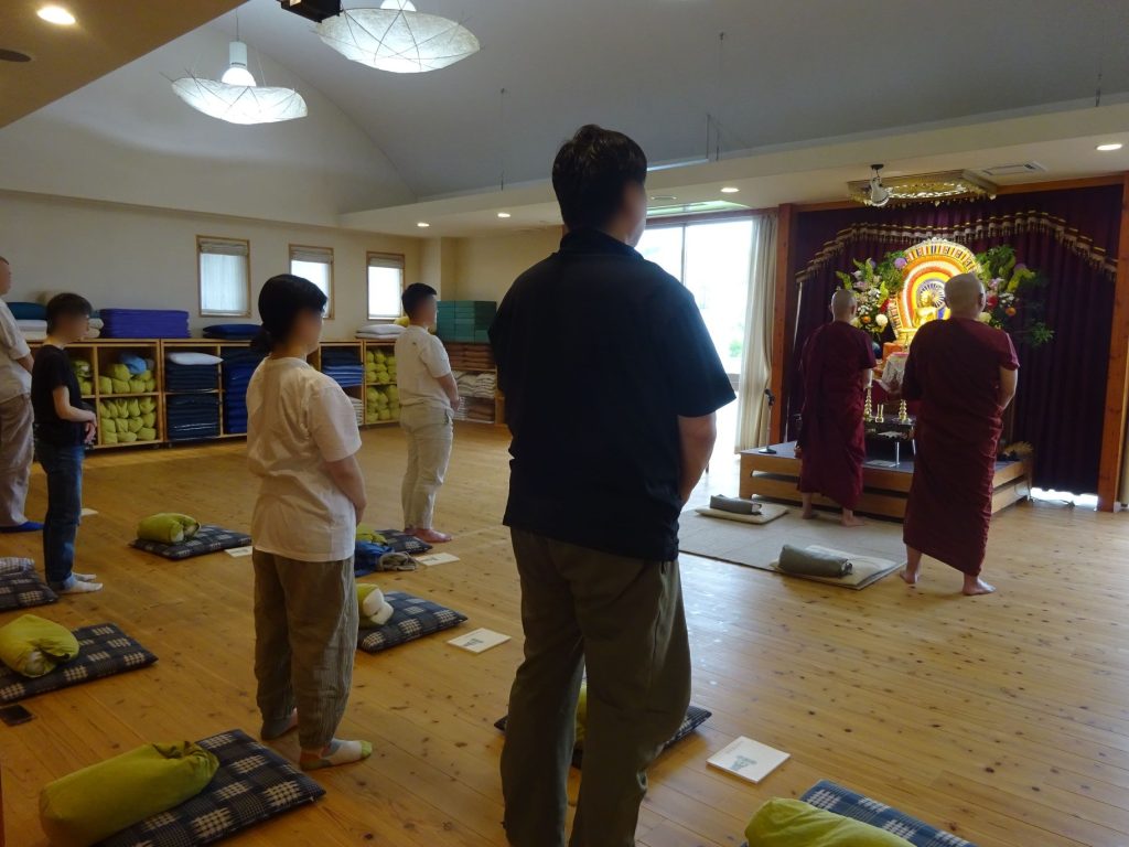 People stand in a meditation hall facing an ornate Buddhist altar with statues and flowers; monks in maroon robes perform a ceremony while others stand on cushions.