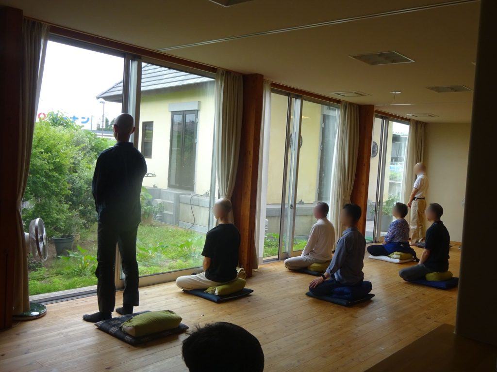 People seated on cushions in a sunlit room, meditating facing large windows with a garden outside.