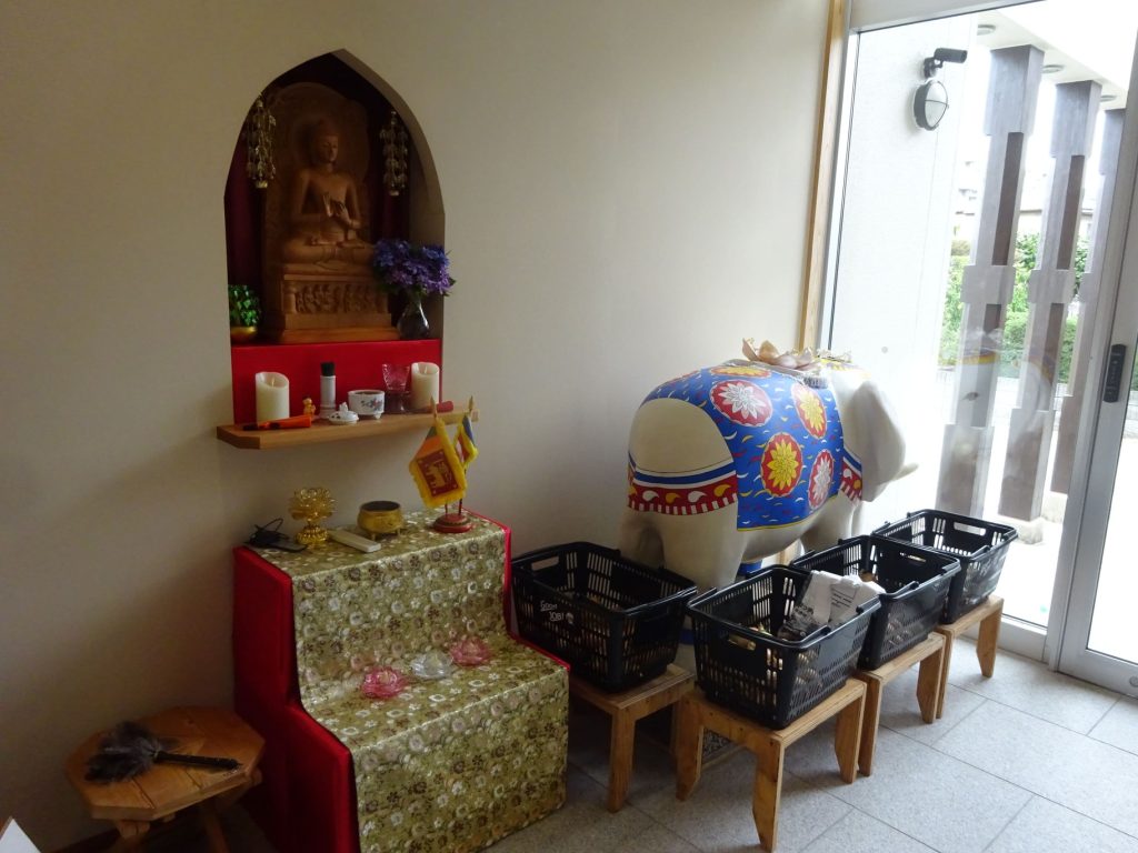 Small Buddhist altar set into a wall niche with a sitting Buddha statue, candles, and flower arrangements on an offering table.