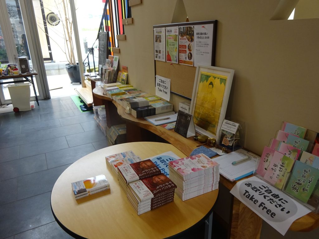 Lobby info desk with pamphlets, brochures, and books on a curved wooden counter; sign reads Take Free in English and Japanese, posters on corkboard behind it.