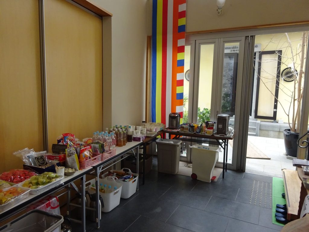 Room with a long table of snacks and drinks along the left wall, colorful banners near glass doors, and a coffee station by the window.