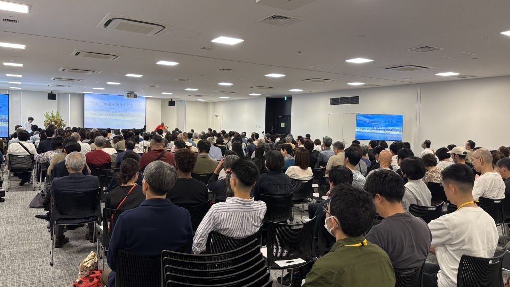 Large conference room filled with a dense audience watching a presenter at the front; two screens show a slide in Japanese.