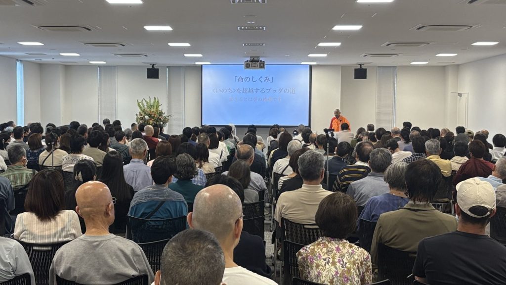 Large audience seated in a conference room, a speaker in orange robes at the podium with a projected screen showing Japanese text in the front center.