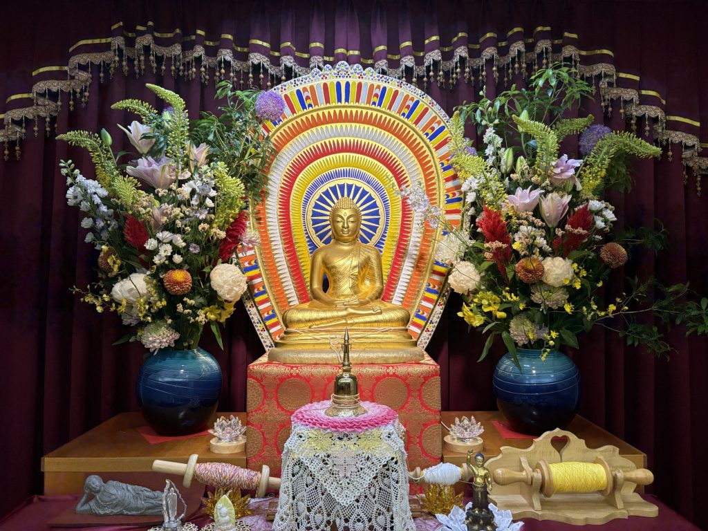 Golden Buddha statue seated in lotus pose on an ornate altar, with a bright rainbow halo behind and lush flower arrangements on either side against a burgundy curtain.