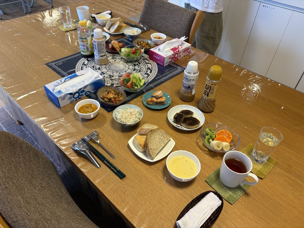 Top-down view of a long wooden table filled with a communal meal: bowls of soup and rice, salads, bread, condiments, fruit, and a tissue box, all set on a plastic-covered table.