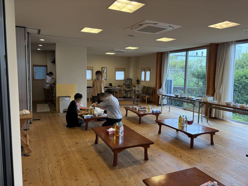Bright community dining room with low wooden tables, people eating, and large windows on the right.