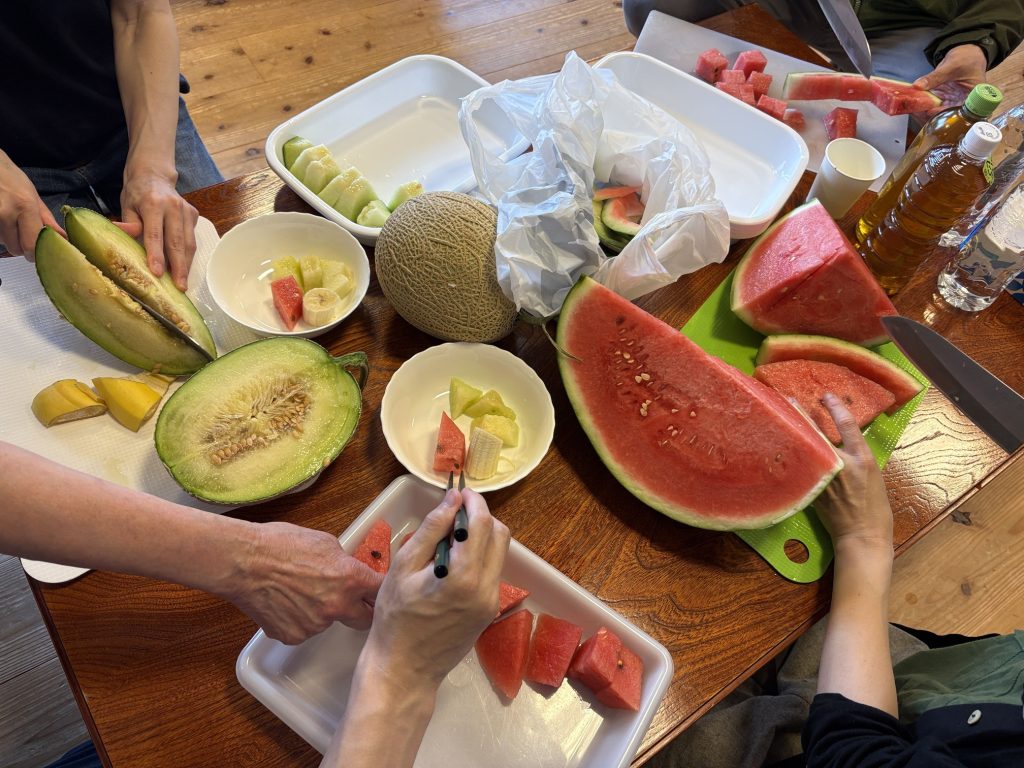 People slicing watermelon and melon on a wooden table, preparing a fruit platter around bowls and cutting boards.