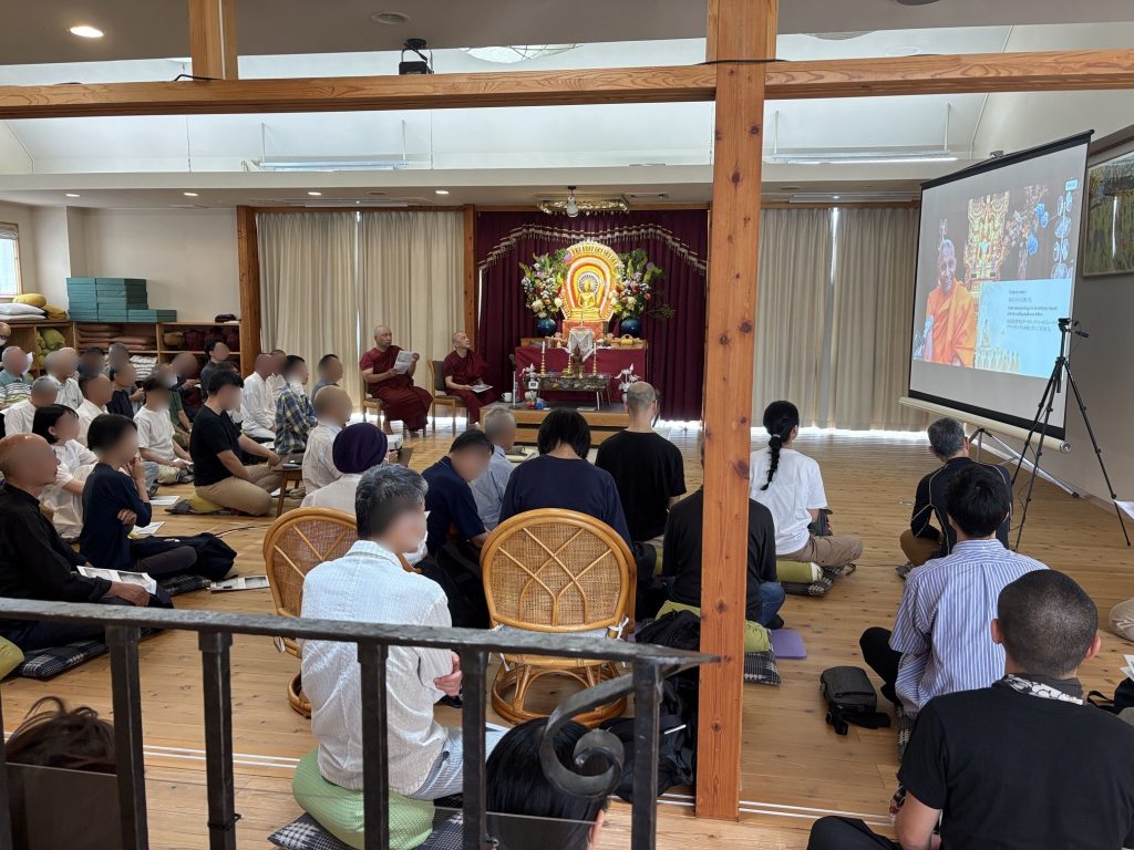 Monks in maroon robes lead a Buddhist ceremony in a hall, with an ornate Buddha shrine and an audience seated on the floor below a projector screen.