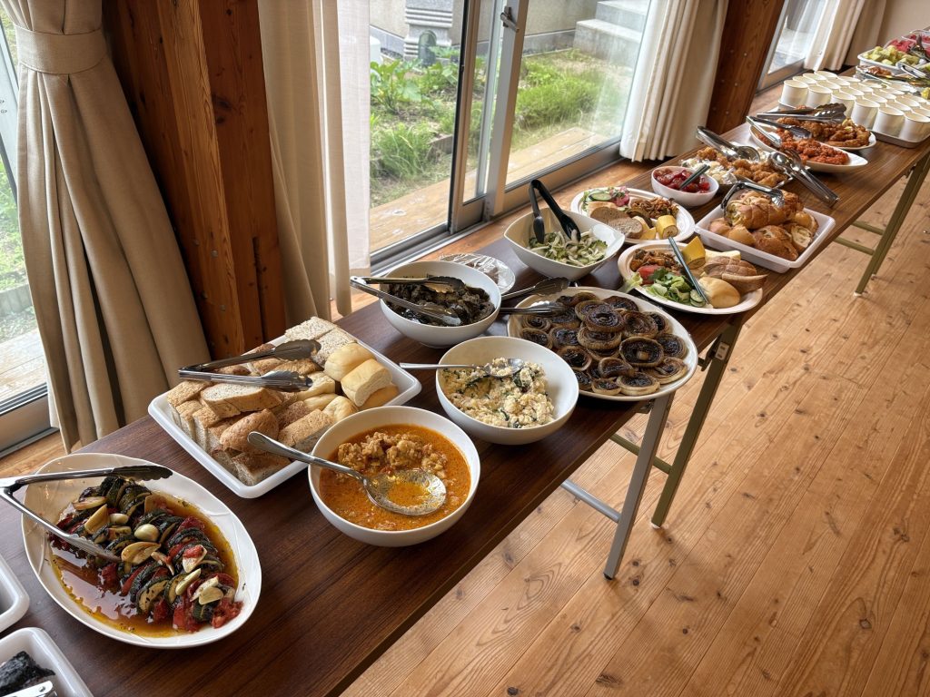 Buffet spread on a wooden table with bowls of salads, breads, roasted vegetables, and pastries near large windows and curtains.