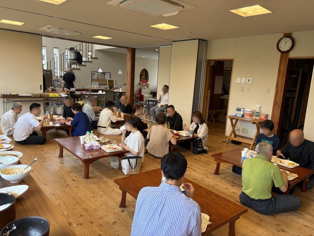 Crowded dining hall with groups of people seated at low wooden tables, eating and drinking; bowls and bottles on the tables.
