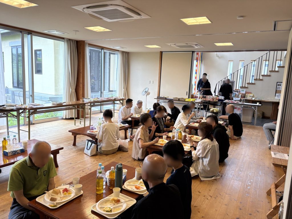 People seated at long tables enjoying a communal meal in a bright room with large windows and a staircase in the background, plates and drinks in front of them.