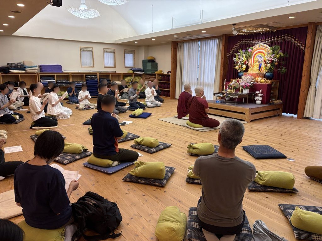 A meditation hall with attendees kneeling on cushions while facing a Buddha altar on a raised platform; monks in maroon robes sit at the front, and shelves of folded mats line the left wall.