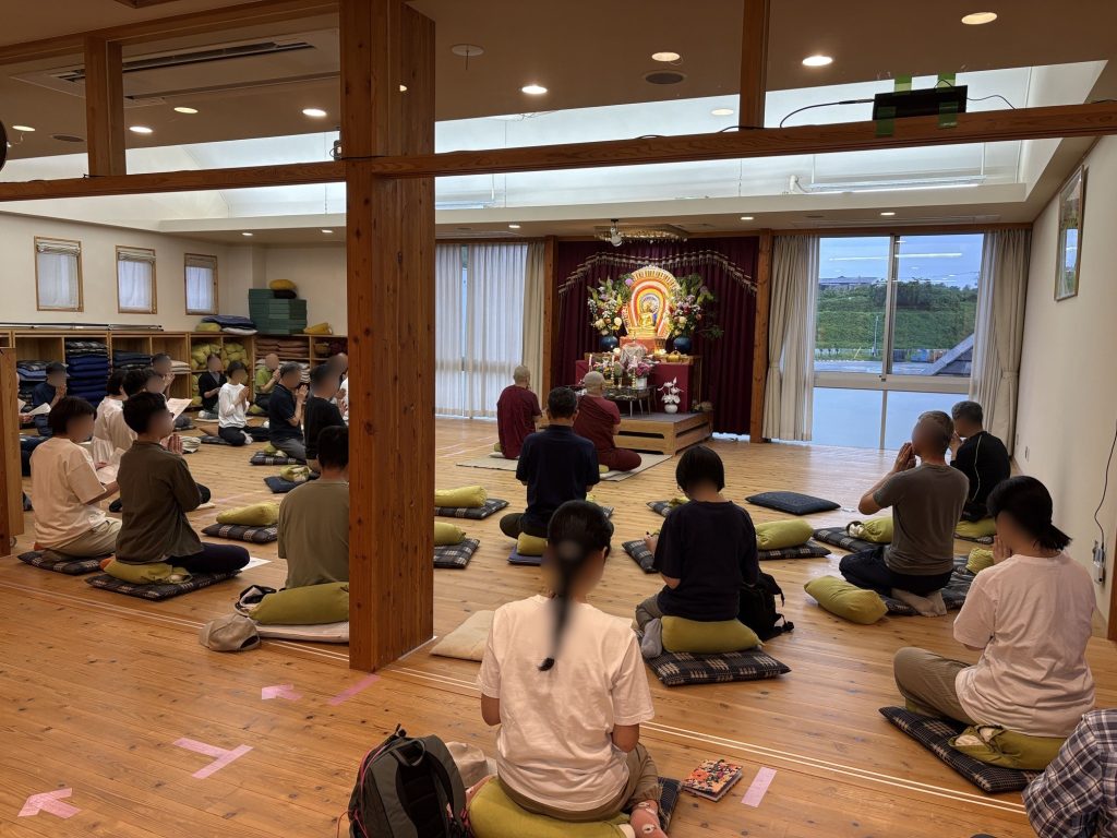 Group of people seated on cushions in a shrine room, facing an ornate Buddha altar with flowers in a bright, wooden interior.