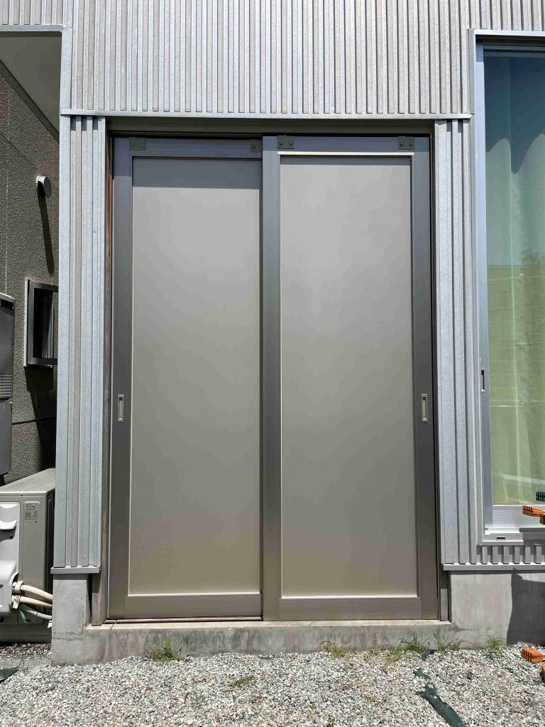 Industrial metal entry door set in a corrugated steel wall, with frosted gray panels and a slim vertical center frame against a concrete base.