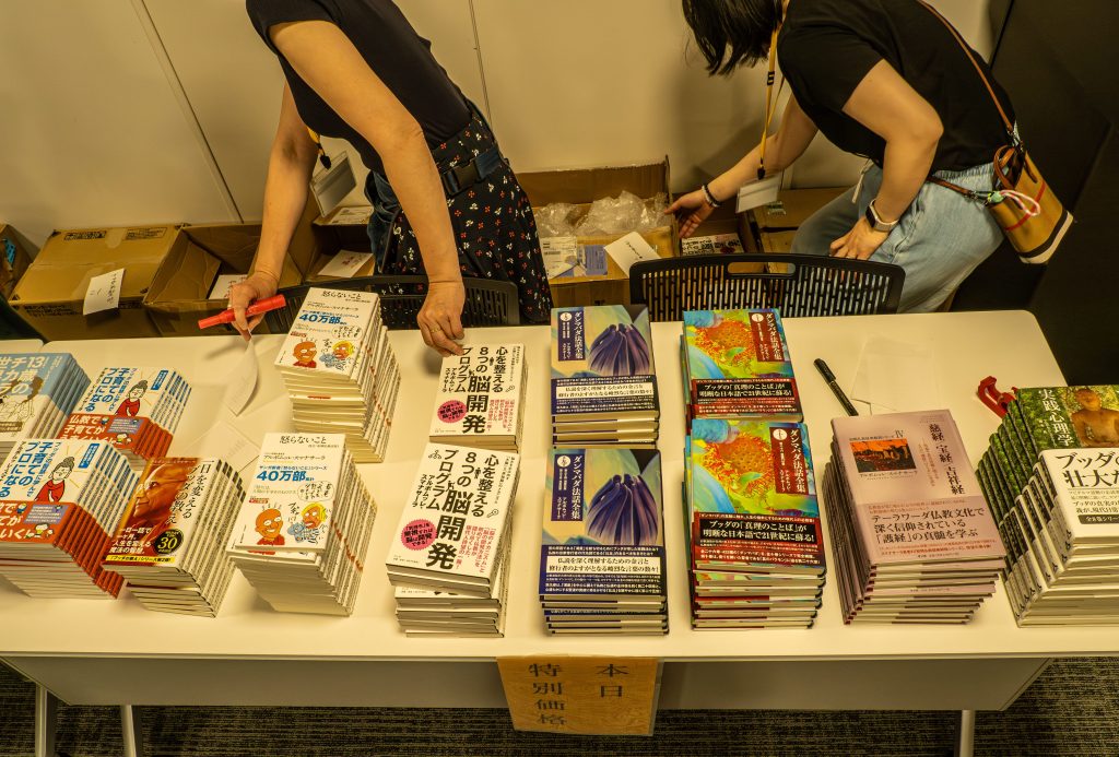 Stacks of Japanese books arranged on a white table at a book sale, with two people browsing in the background.
