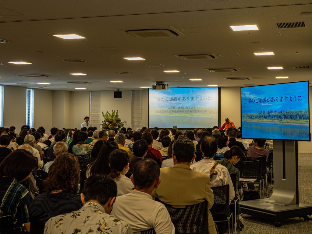 Crowded conference room with attendees facing two large screens displaying Japanese text.