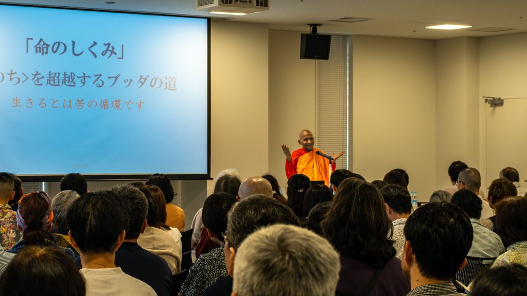 Buddhist monk in orange robes giving a talk at a podium in a conference room with a projection screen on the left.