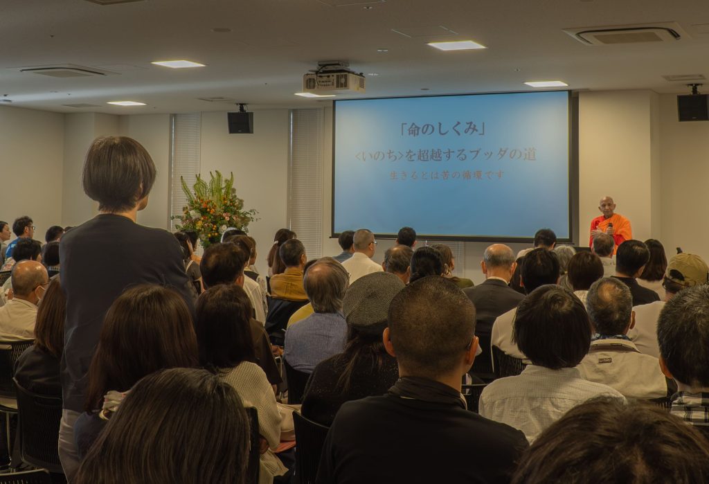 Audience seated in a conference room facing a large screen with Japanese text; a monk in orange robes stands at right front.