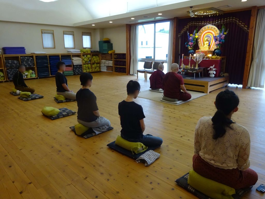 Meditation session in a Buddhist hall with laypeople and monks seated on cushions facing a Buddha altar.