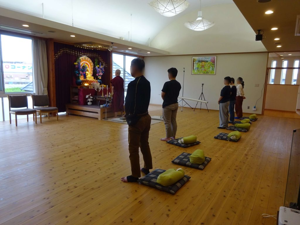 People standing in a meditation room facing a Buddha altar with flowers and candles on a stage.