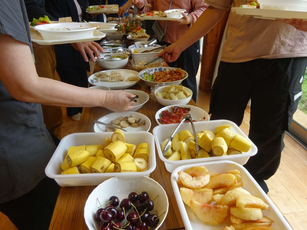 People at a buffet select food from a long table of bowls and plates, with bowls of cherries, bananas, and other fruits visible.