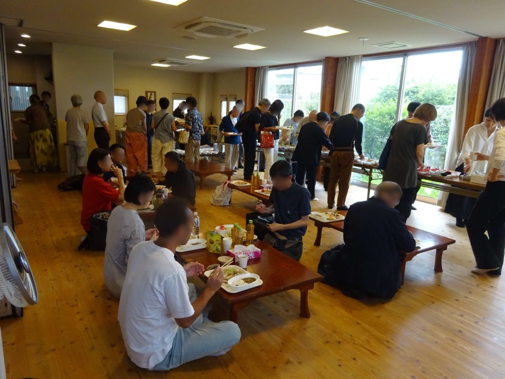 Crowded dining room with people lining up for buffet and others seated at tables enjoying meals at a gathering.