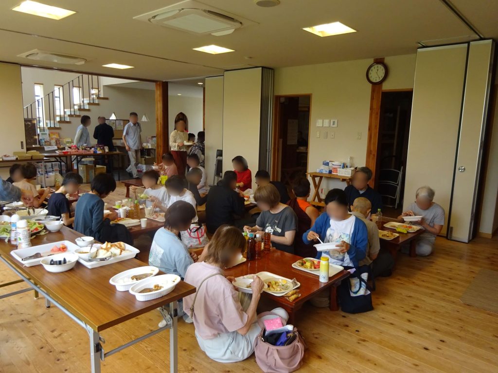 Large group of people seated at long wooden tables, eating meals in a bright dining hall.