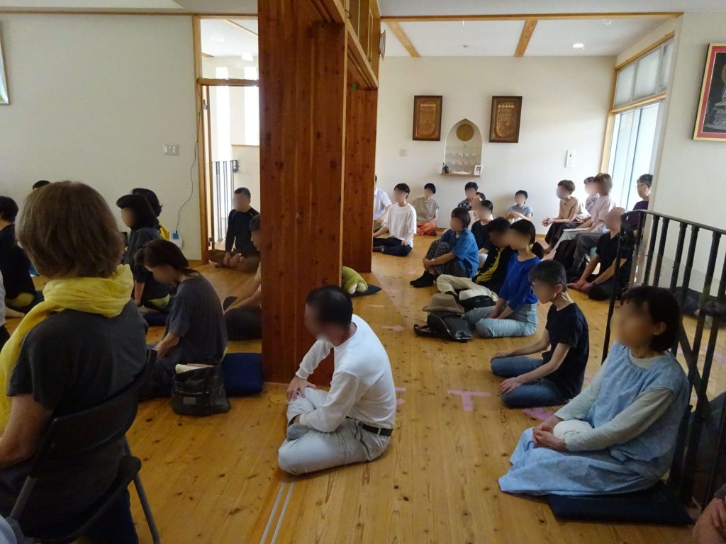 People sit on the wooden floor of a bright room in a group session, arranged around a central wooden pillar, listening attentively.