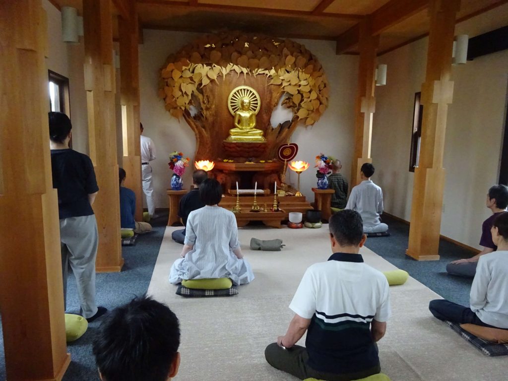 Prayer hall with a golden Buddha statue in a wooden shrine, surrounded by warm lights and flower offerings; people sit kneeling on cushions in meditation.