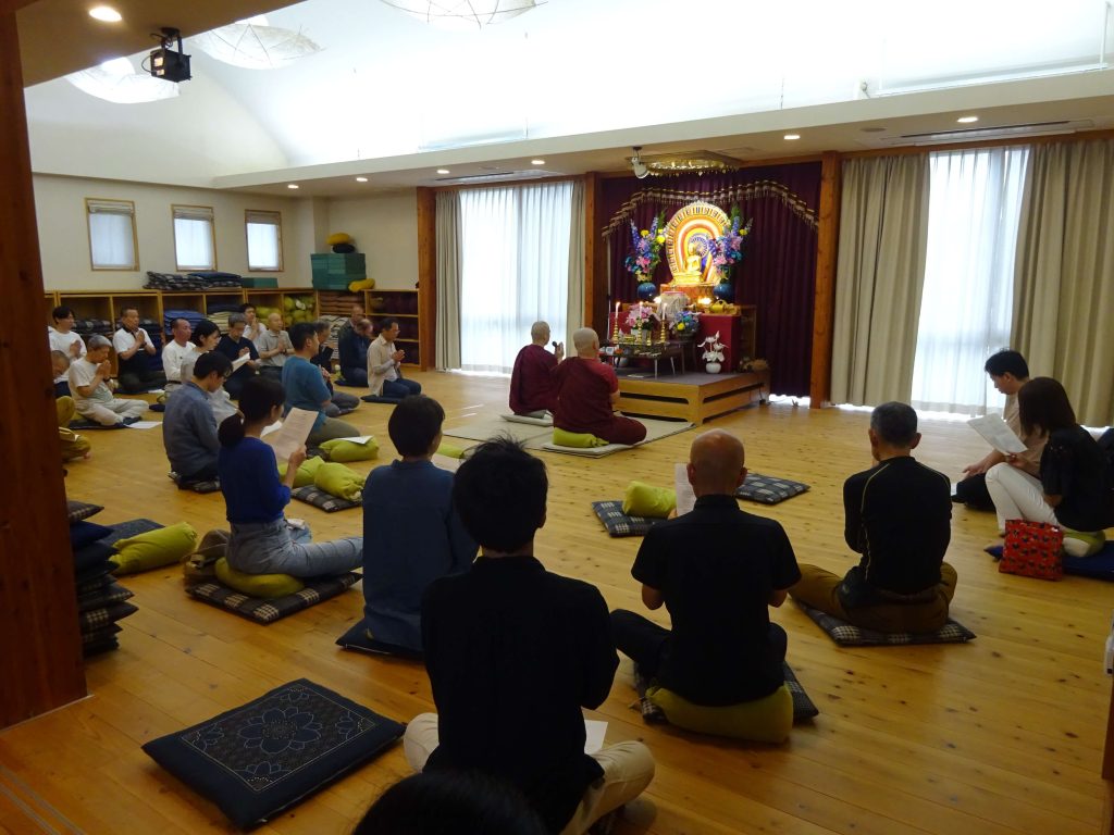 Group of people seated on cushions in a bright meditation hall facing a Buddhist altar with a statue and floral arrangements.