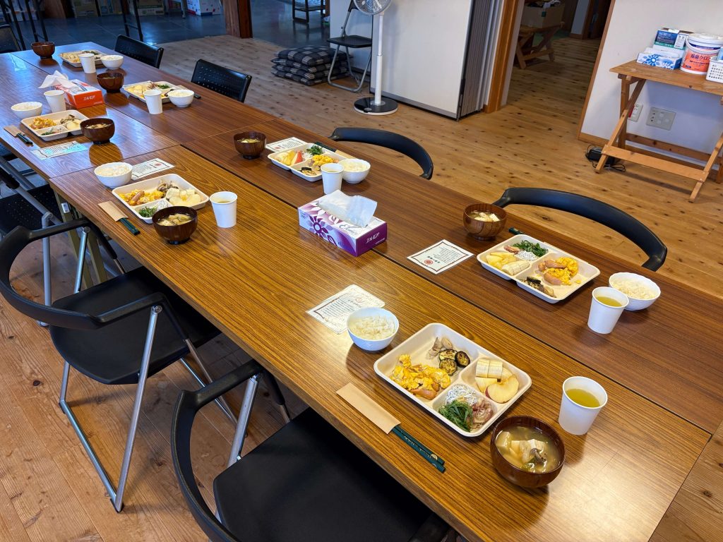 Long wooden table set with multiple bento-style trays, bowls of soup, paper cups, and chopsticks in a casual dining room in an office or community space.