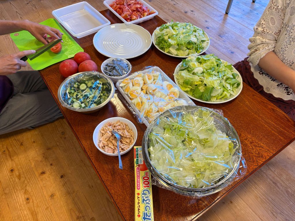 Person chopping a tomato on a bright green cutting board with bowls of salad ingredients (lettuce, tomatoes, cucumbers) on a wooden table.