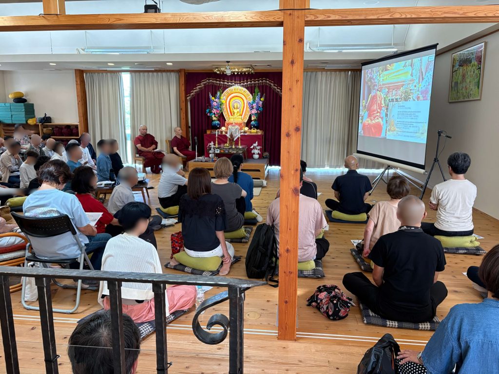 Meditation hall with monks in maroon robes facing a seated audience, shrine altar at center and a projector screen on the right.