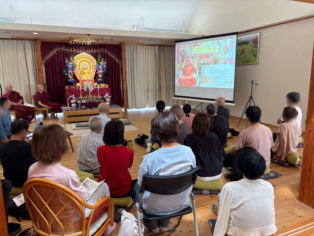 Indoor meditation hall with seated monks and an audience watching a projected slide beside an ornate altar and Buddha statue with flowers and candles.