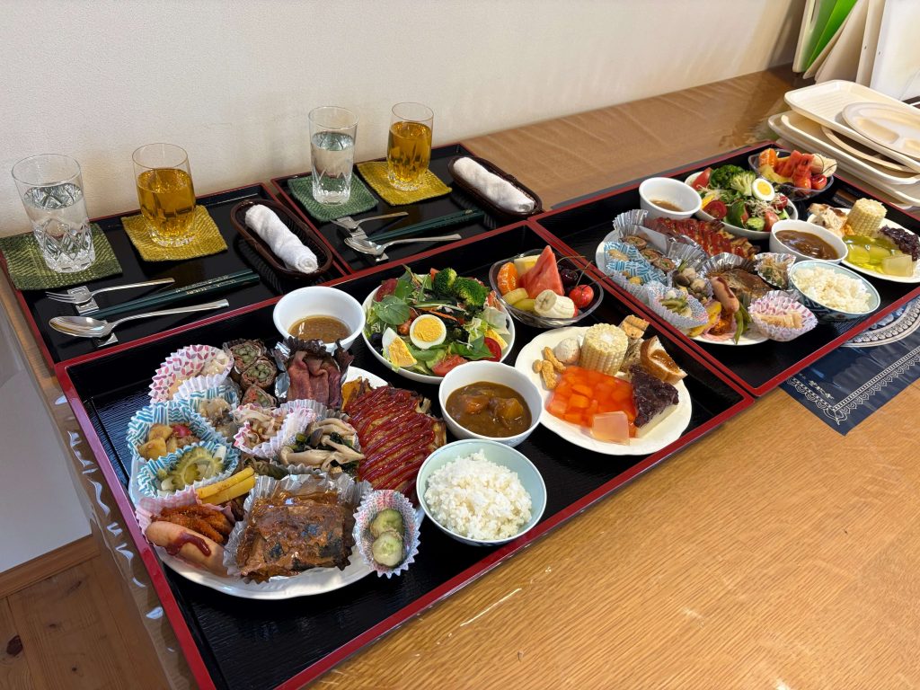 Large multi-tier Japanese bento spread on black lacquer trays with assorted dishes, rice bowls, salads, and skewers on a wooden table.