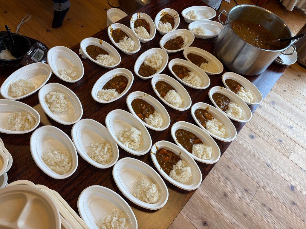 Rows of white oval bowls filled with curry and rice on a wooden table, ready for serving; a large pot of curry on the right.