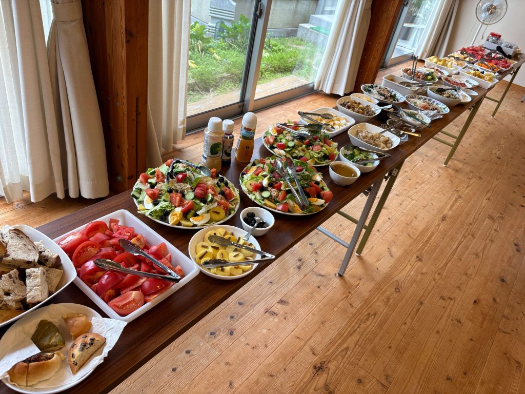 Buffet table with bowls of salad, fruit, and breads along a wooden floor in a bright room with large windows.
