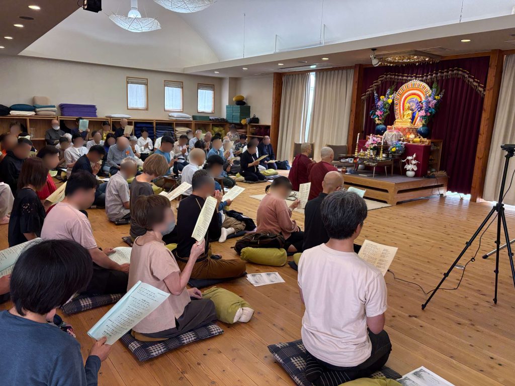 Group of people kneeling and sitting on cushions in a temple hall, holding prayer sheets near an altar with a Buddha statue and flowers on the right.