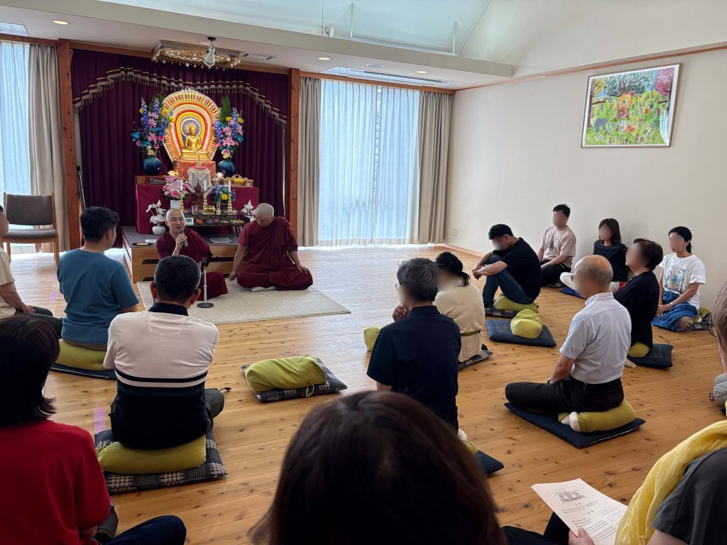 Meditation hall with a gold Buddha statue on an altar, monks speak to a seated audience on cushions in front of a burgundy curtain backdrop.