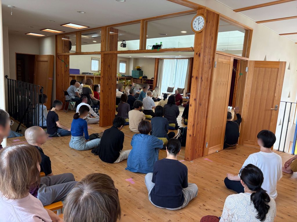 People sit cross-legged on cushions in a wooden meditation hall, listening to a monk at the front.