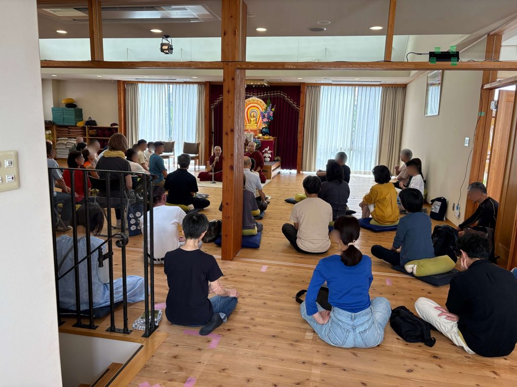 Monks leading a meditation talk in a bright room; a Buddha altar and flowers at the front while attendees sit on cushions on a wooden floor.