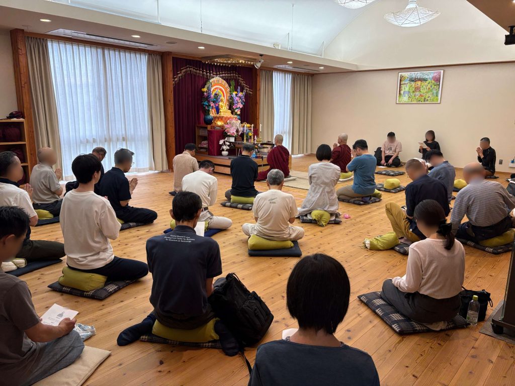 Group of people kneeling on cushions in a bright room, facing a colorful altar with a Buddha statue and flowers for a meditation or prayer ceremony.