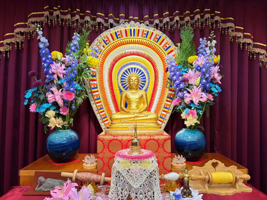 Golden Buddha statue seated in meditation on an ornate shrine, with a vibrant rainbow mandala behind and colorful flowers on both sides against burgundy curtains.
